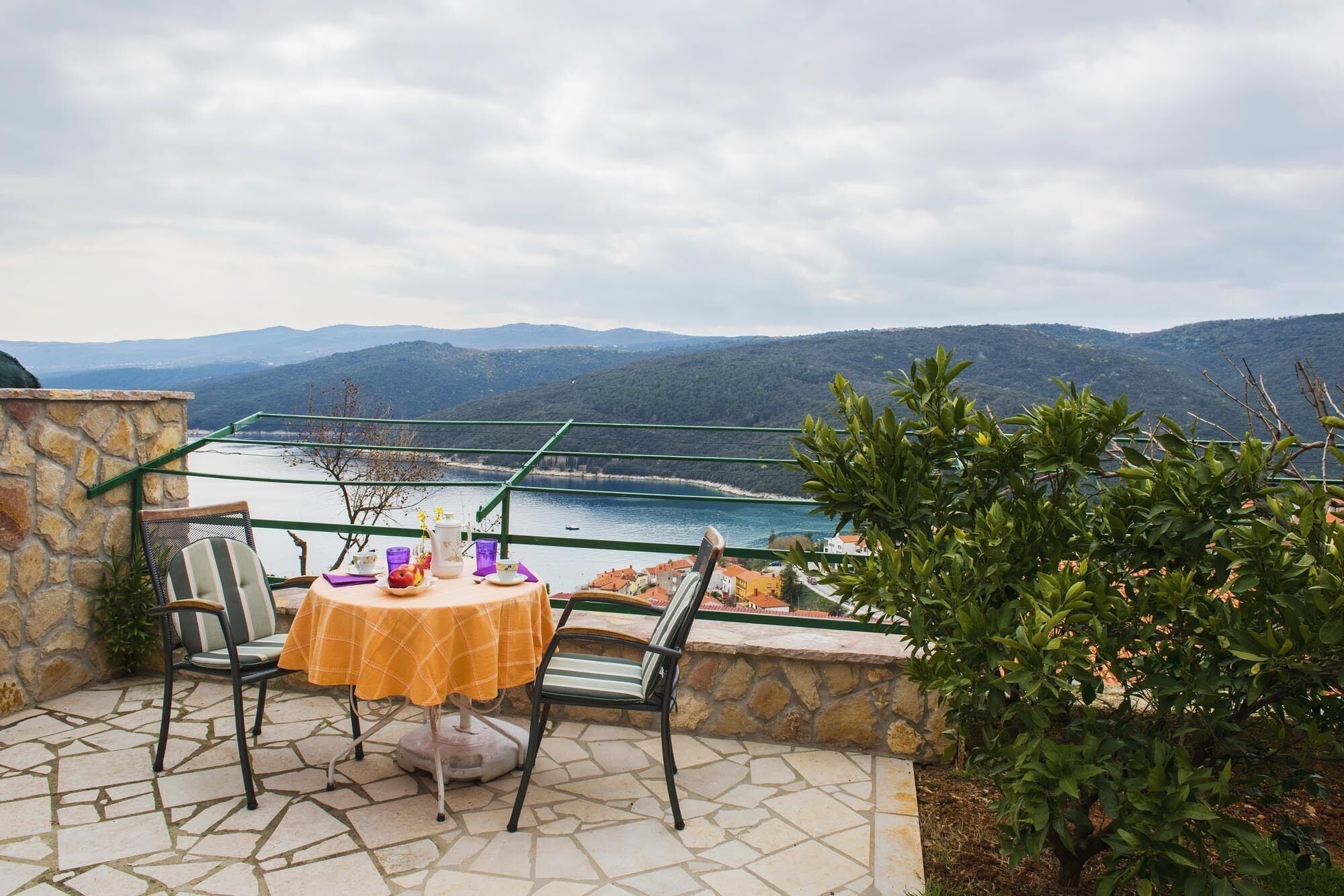 A terrace with a table set for two, overlooking a body of water and hills.