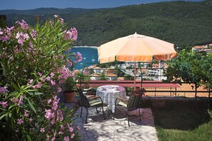 A patio with a table and chairs, an umbrella, and a view of the sea and buildings in the background. - Apartment 2023/28734 (Istria - Rabac), 500m from the beach (Rabac)