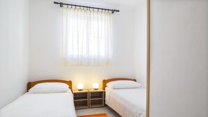 A small, clean room with two single beds, a nightstand with a lamp, a window with sheer curtains, and a wooden floor.
