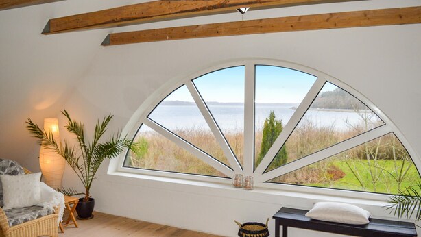 Ein Zimmer mit einem großen Bogenfenster, das einen Blick auf einen See und viel Grün bietet. Es gibt ein Korbsessel, einen kleinen Tisch mit Dekoration und eine Bank mit Kissen.