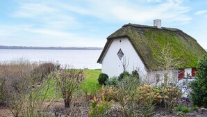 Ein weißes Haus mit Strohdach, einem kleinen Garten und Blick auf ein Gewässer.
