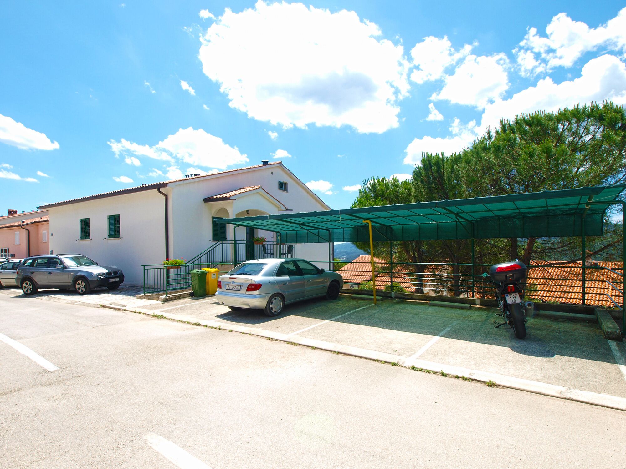 A parking area with a covered motorcycle stand, a car, and a building with a tiled roof.