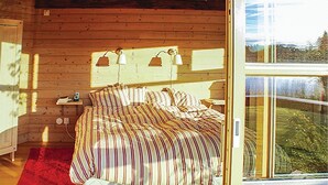 Ett mysigt sovrum med ett träpanelat tak, en stor säng med randiga sängkläder, två sänglampor och en utsikt över grönskan genom ett stort fönster.