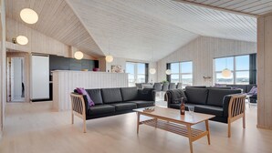 A spacious living area with a high ceiling, wooden walls, and large windows offering a view of the outdoors. - Nice home in Løkken with sauna (Løkken)