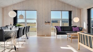 A spacious room with a high ceiling and wooden walls, featuring a dining area with a checkered tablecloth, a black leather sofa, and a small wooden coffee table. - Nice home in Løkken with sauna (Løkken)
