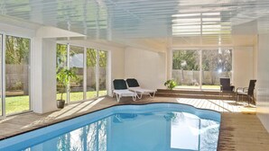 Une piscine intérieure avec des parois en verre transparent, une terrasse en bois et des chaises longues.
