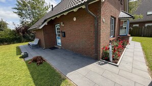 A brick house with a tiled roof, a front porch with a bench, and a dog lying on the grass. - Vacation home Queller 14, fenced garden, Wlan - Holiday home Queller 14 (Friedrichskoog)