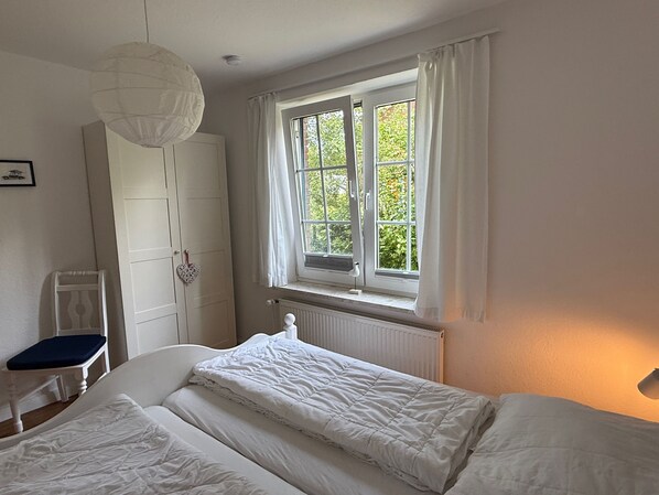 Quarto com cama, cadeira, guarda-roupa, janela com cortinas e radiador.