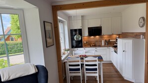 A modern kitchen with white cabinets, a wooden ceiling, and a dining area with a table and chairs. - Vacation home Queller 14, fenced garden, Wlan - Holiday home Queller 14 (Friedrichskoog)