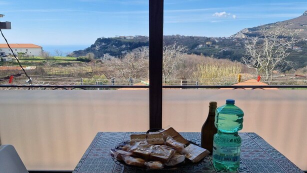 Une table avec du pain et une bouteille d’eau sur un balcon offrant une vue sur des montagnes et une étendue d’eau.
