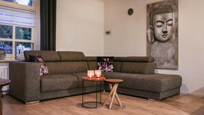 A living room with a grey sectional sofa, two small wooden tables, a Buddha painting, and a window with curtains.