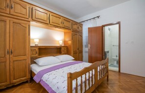 Une chambre à coucher avec un lit en bois, une table de chevet et une armoire. On peut apercevoir la salle de bain à travers une porte en verre.