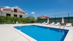 A rectangular swimming pool with a clear blue water surface, surrounded by a tiled deck. There are white lounge chairs and umbrellas arranged for sunbathing. A fence encloses the area, and a building with a balcony is visible in the background. - Amazing home in Runovic (Runovic)