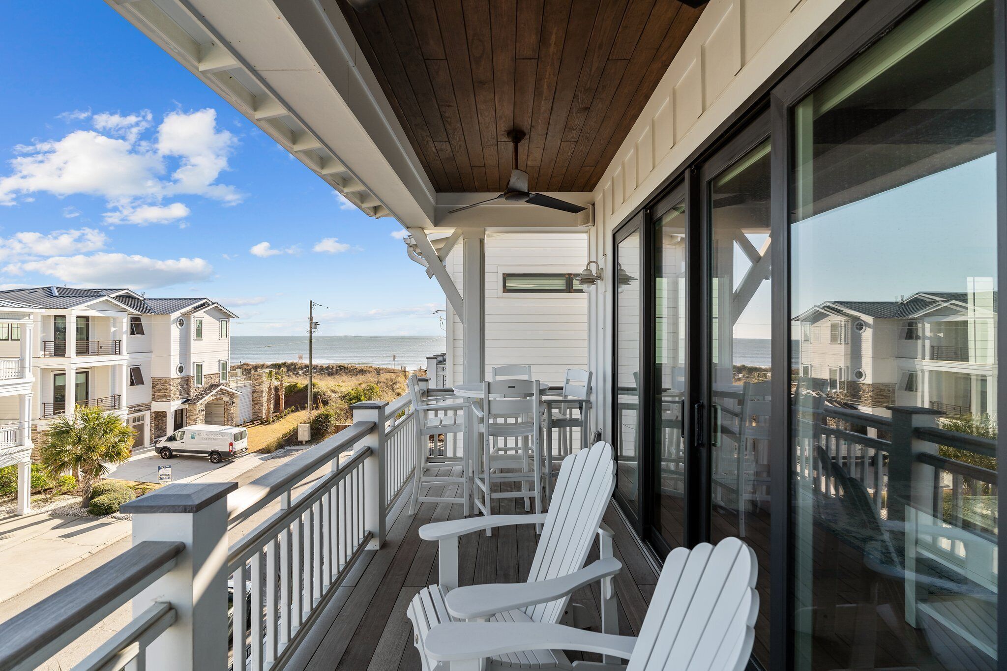 A balcony with white Adirondack chairs, a ceiling fan, and a view of the beach and ocean.