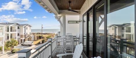 A balcony with white Adirondack chairs, a ceiling fan, and a view of the beach and ocean.