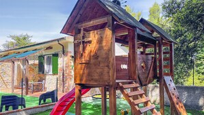 Une cabane en bois avec un toboggan et une échelle d’escalade, entourée d’une zone délimitée par une clôture et dotée d’un espace extérieur pour s’asseoir.