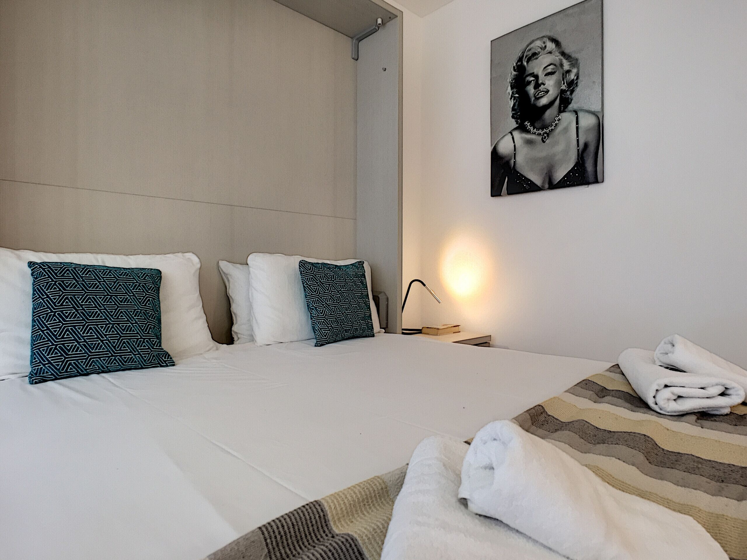 A hotel room with a large bed, two patterned pillows, a framed picture on the wall, a bedside lamp, and a rolled towel on the bed.