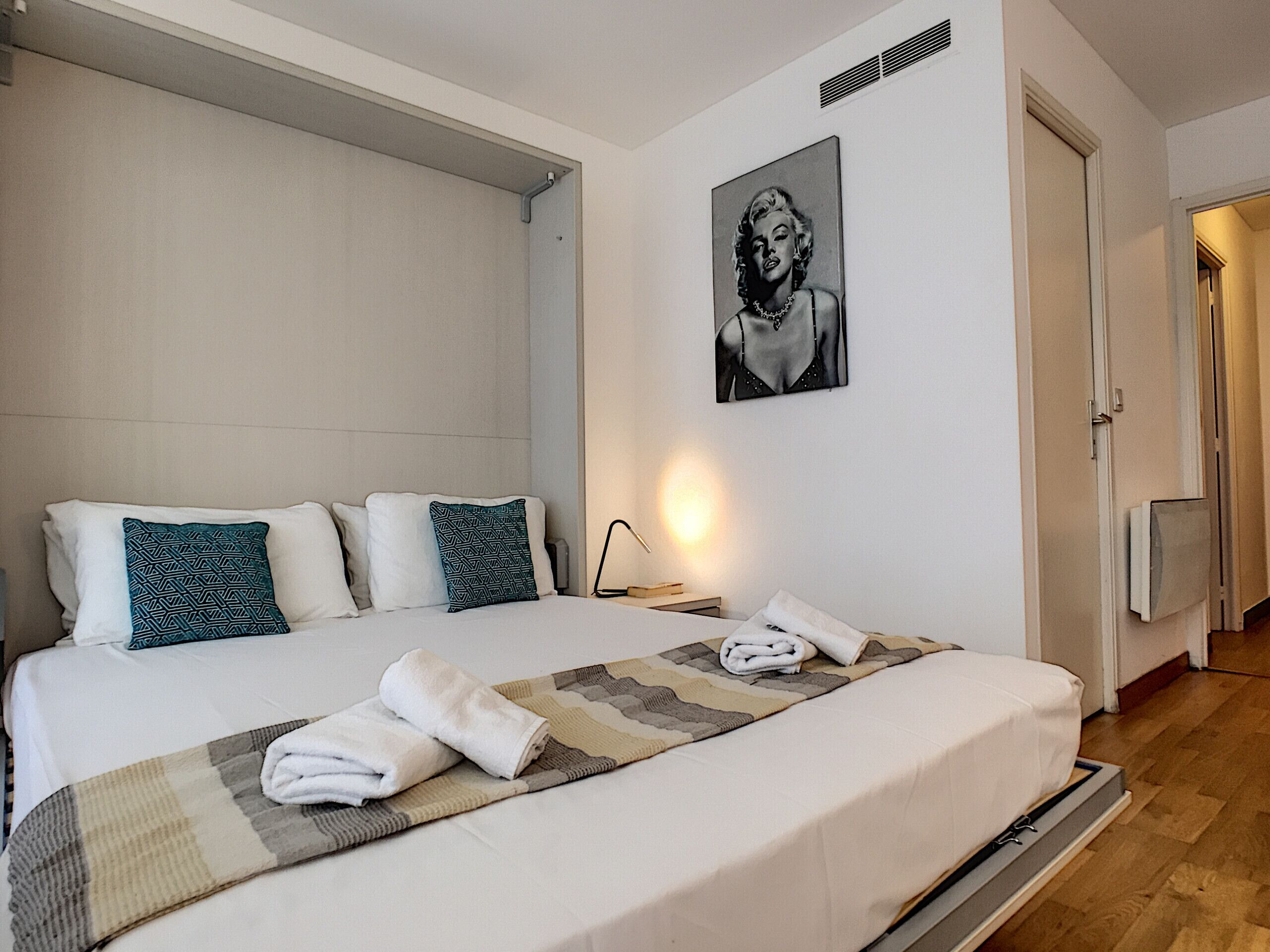 A neatly made bed with pillows, a blanket, and towels. There is a painting on the wall and a lamp on the bedside table.