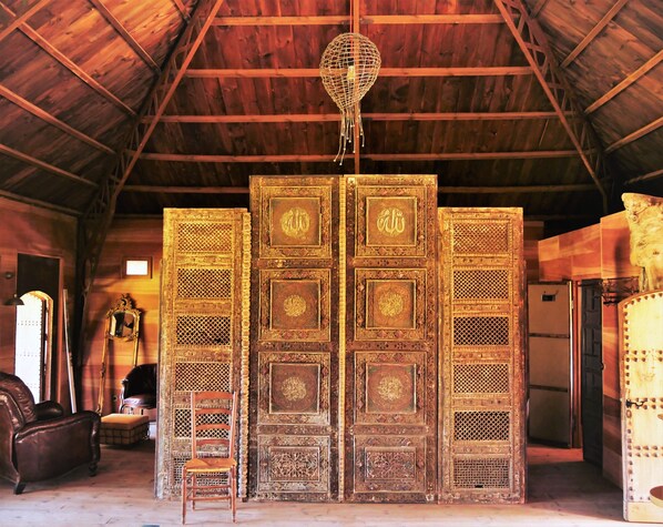 A room with a high wooden ceiling, a large ornate door with religious inscriptions, a hanging light fixture, a leather sofa, and a wooden chair.