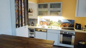 Una cocina compacta con armarios blancos, horno empotrado y cocina. Se aprecia una ventana con vistas a zonas verdes y un cuadro sobre la encimera.