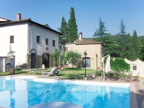 Une piscine avec des chaises longues, un bâtiment blanc aux murs en pierre et un jardin avec des arbres.