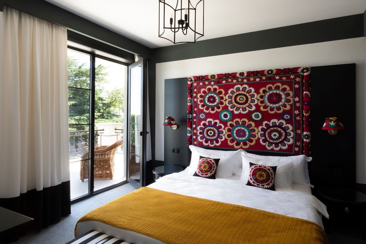 A modern hotel room with a large bed, a view through a sliding glass door, and a patterned tapestry on the headboard.