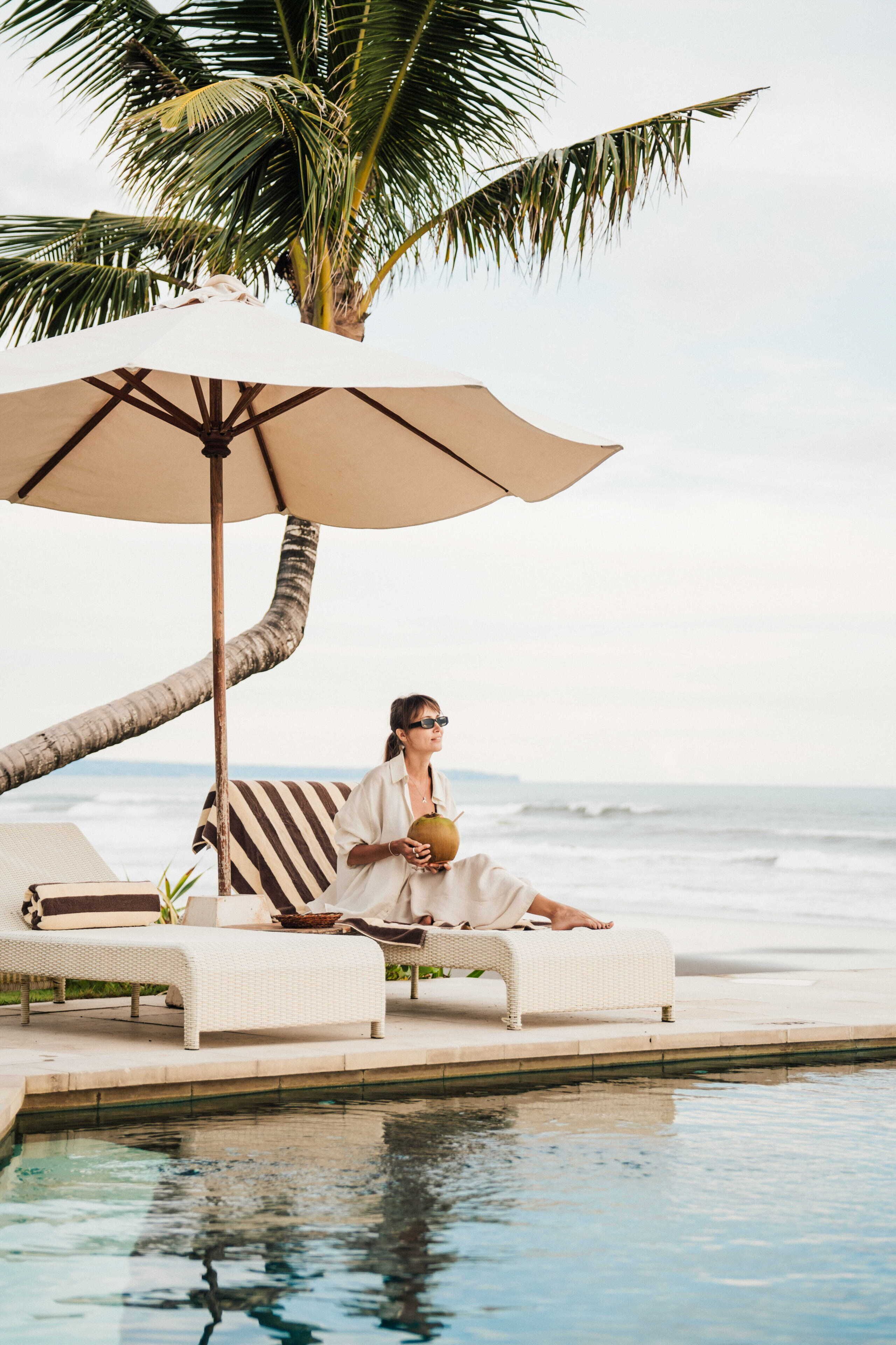 En person slapper af i en hvid liggestol ved en pool med palmetræ og havudsigt.