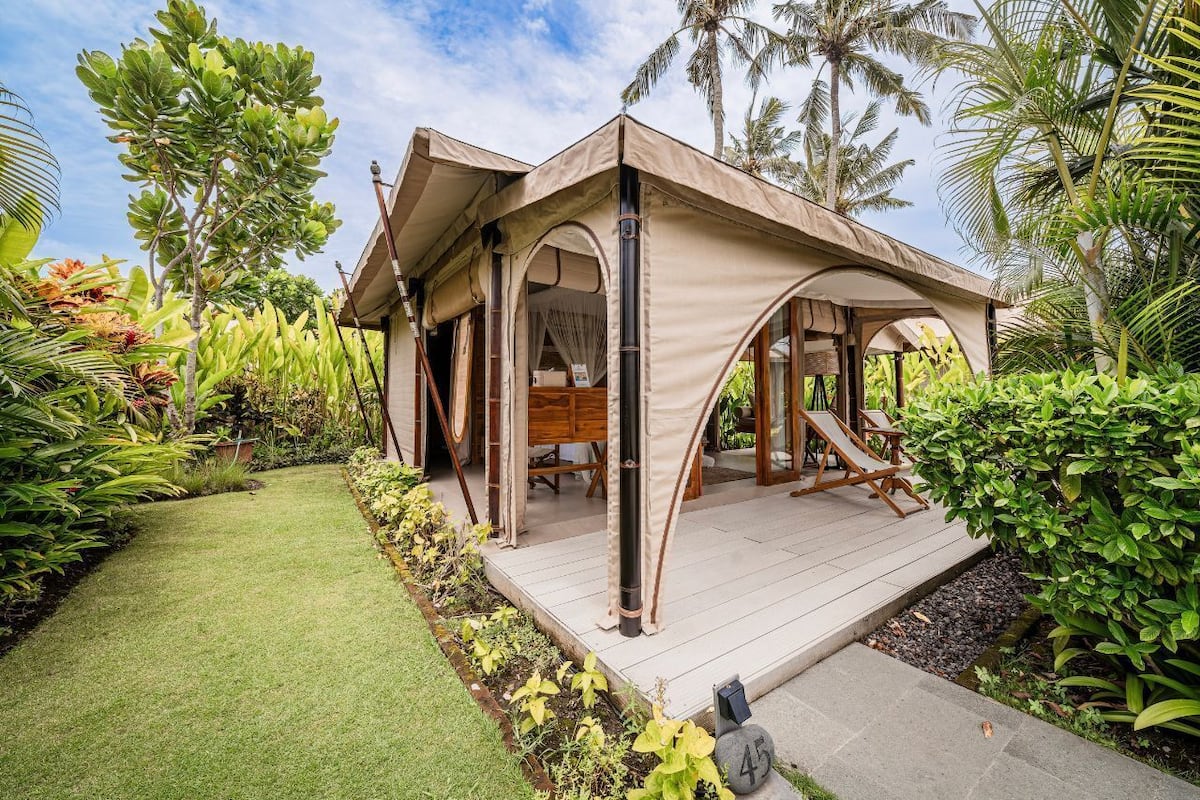 A tent-style accommodation with a deck, surrounded by lush greenery and palm trees.