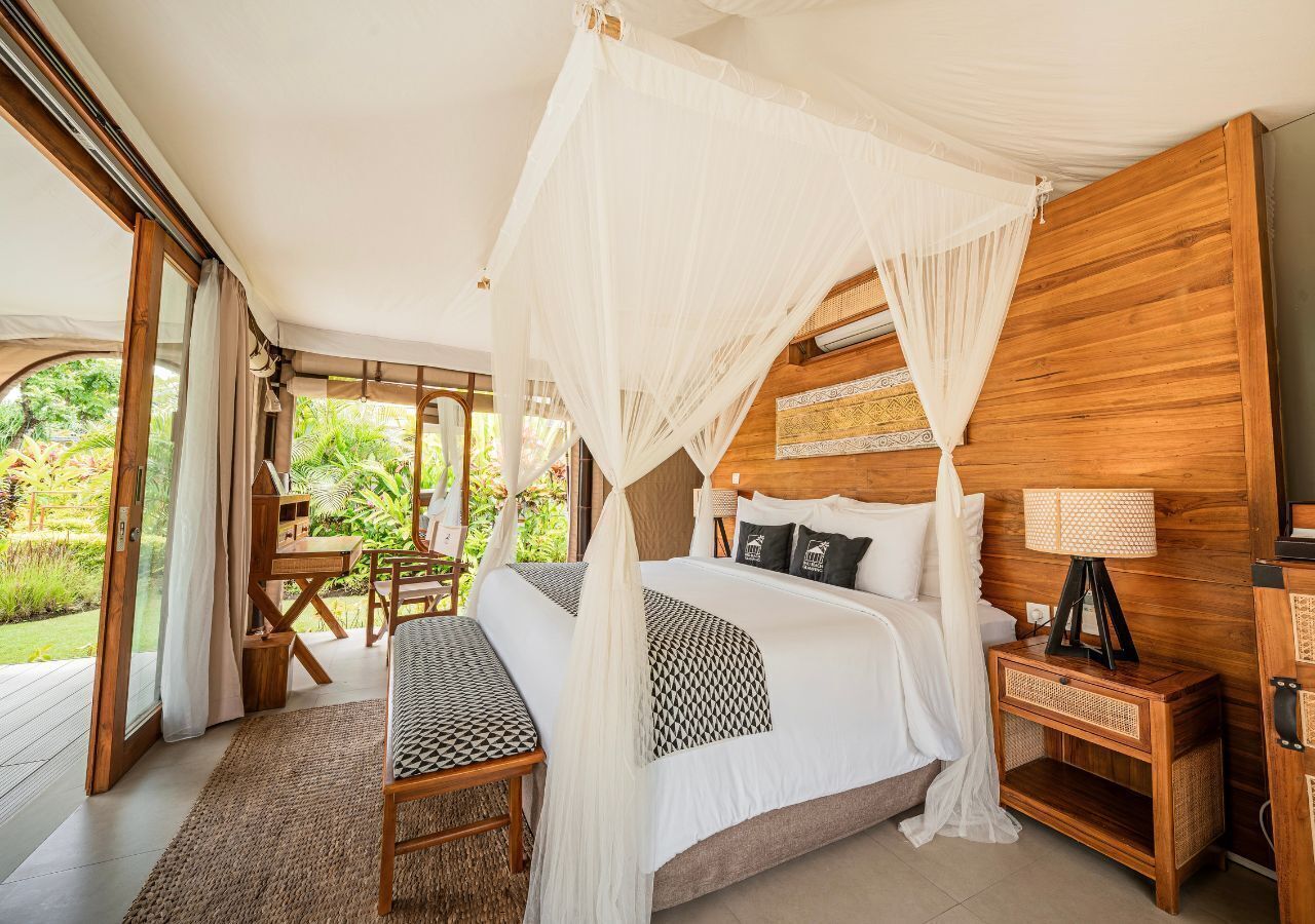 A bedroom with a canopy bed, wooden headboard, bedside table with lamp, and a view of greenery outside.