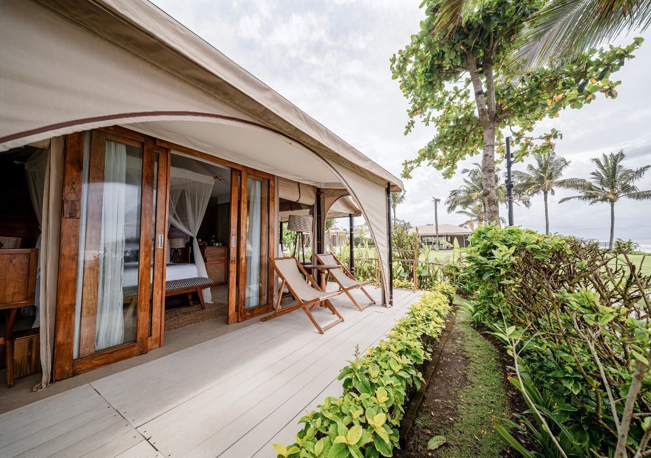 Een villabehuizing aan het strand met een overdekt terras, houten meubilair en weelderige begroeiing.