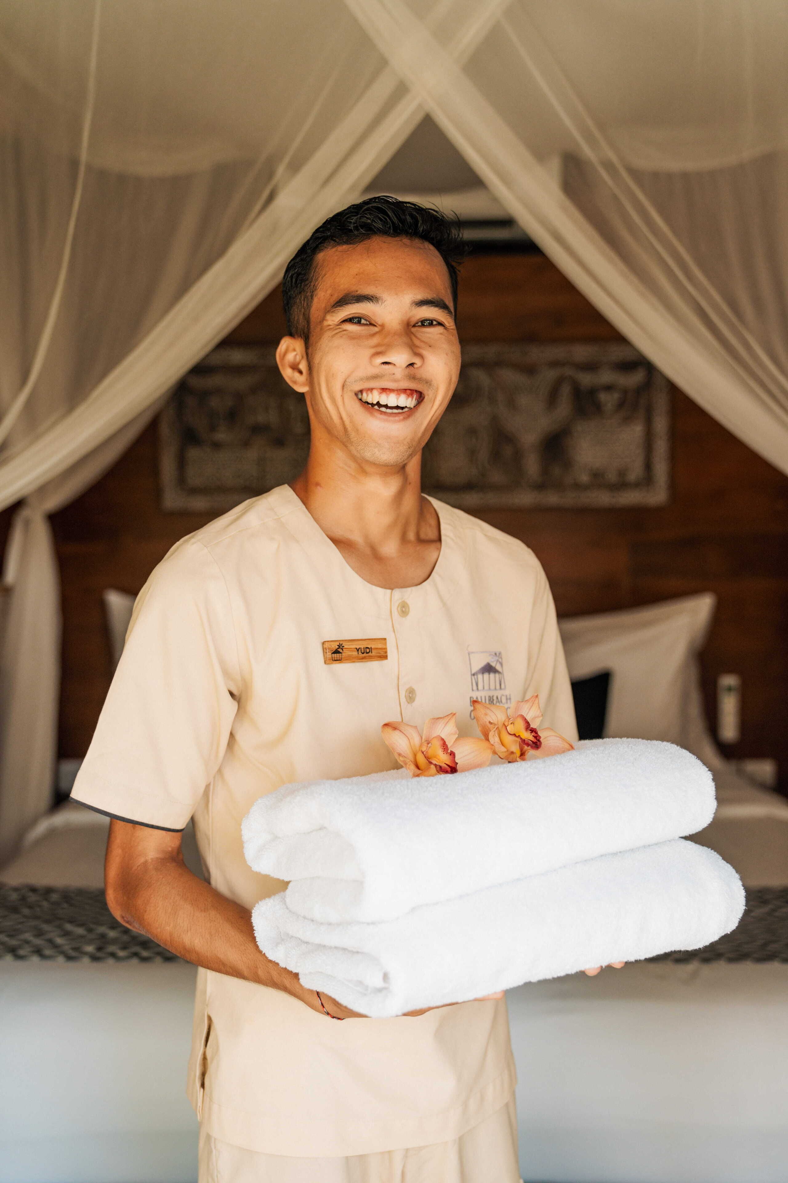Eine Person hält gefaltete weiße Handtücher in einem Raum mit einem Bett, einem Baldachin und einem an der Wand hängenden Bild.