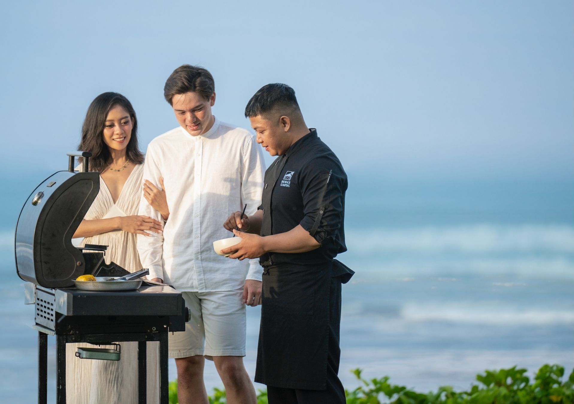 Trois personnes sont réunies autour d’un barbecue, avec une vue sur l’océan.