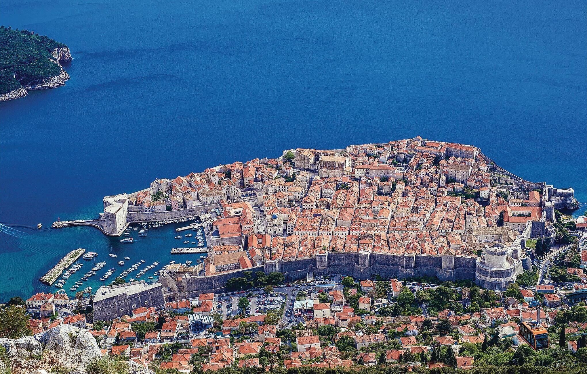 An aerial view of a coastal town with a dense cluster of buildings, a fortified wall, and a marina with boats.