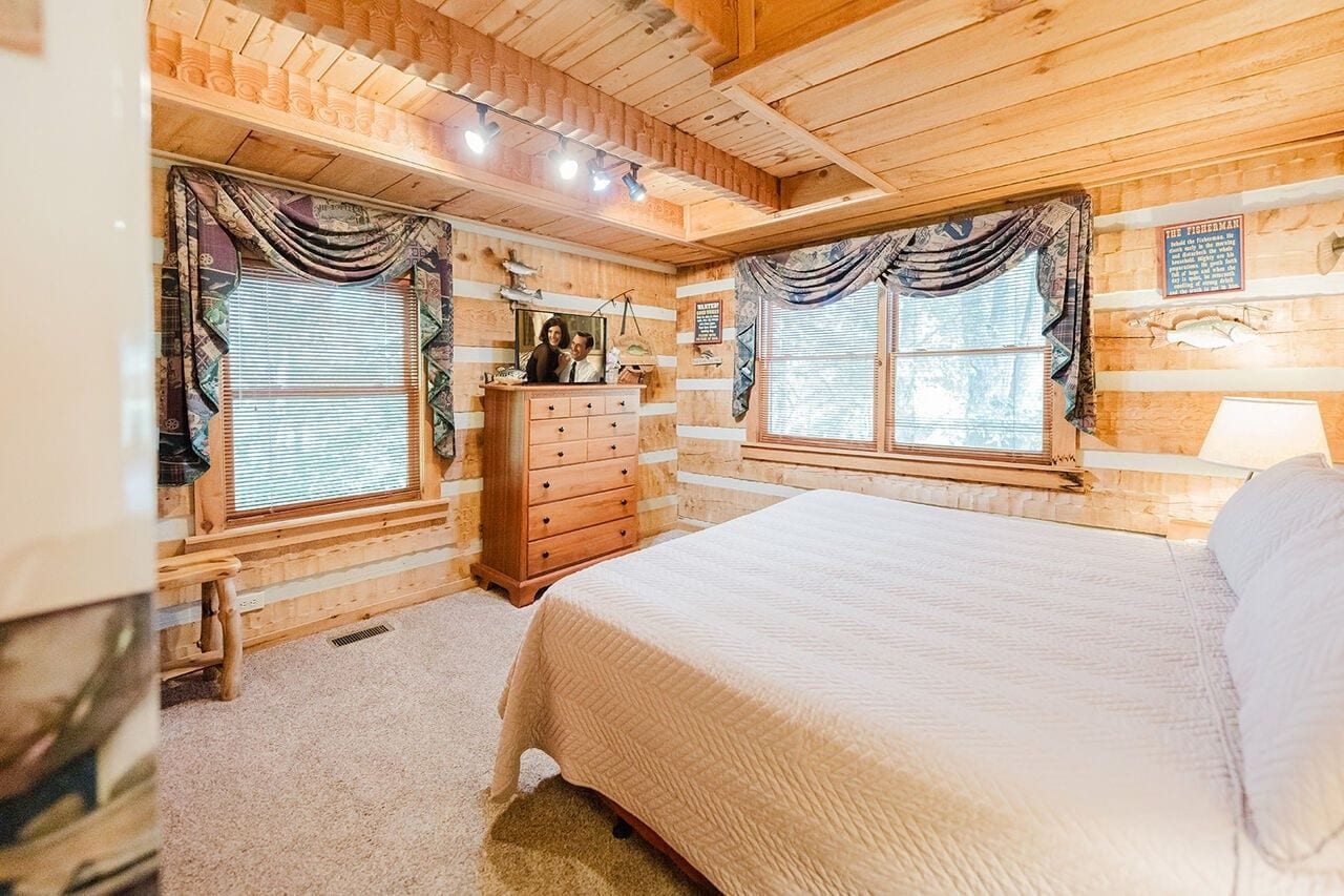 A wooden cabin bedroom with a bed, dresser, and two windows with curtains.