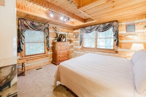Un dormitorio de cabaña de madera con cama, cómoda y dos ventanas con cortinas.