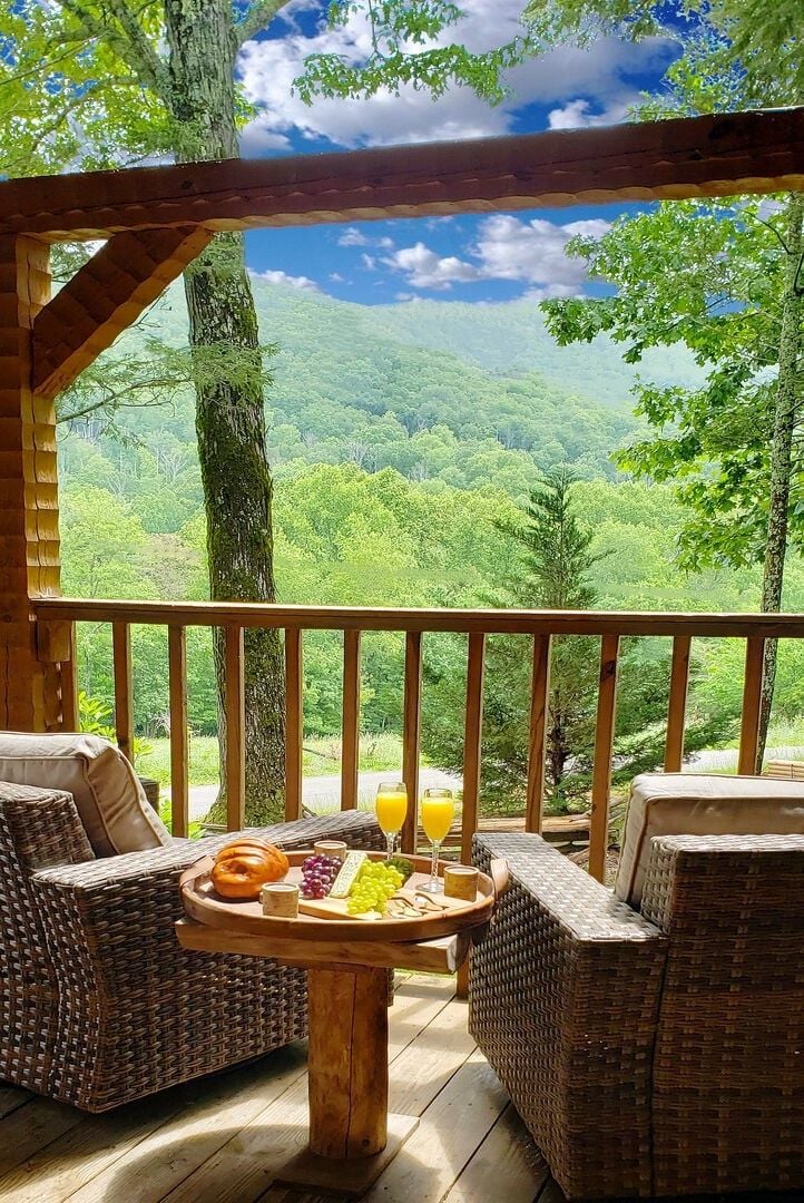 Terraza de madera con mobiliario de mimbre y una mesa dispuesta con comida y bebidas, con vistas a un paisaje boscoso.
