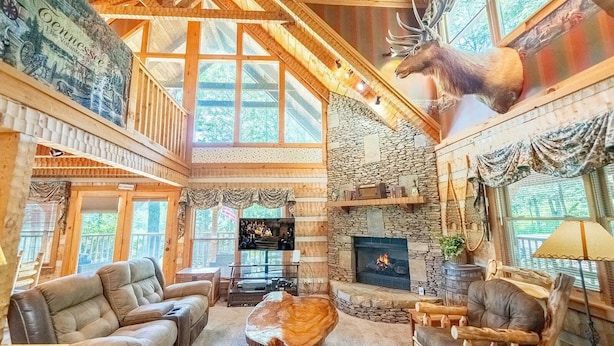 A rustic cabin interior with a stone fireplace, leather furniture, and a mounted deer head. - Big Kahuna Lodge 3 Bedroom Cabin by RedAwning (Sevierville)