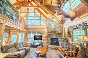 A spacious lodge interior with a stone fireplace, wooden furniture, and a mounted elk head.