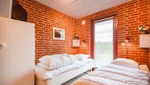 Une chambre avec un mur en briques, un lit, une fenêtre avec des stores et un petit meuble.