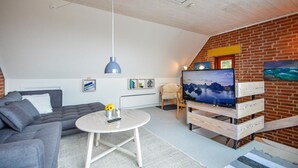 Un salon moderne avec un canapé gris, une table basse blanche, un téléviseur posé sur un meuble en bois et un mur en briques.