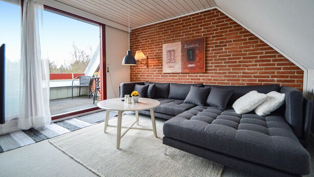 Un salon moderne avec un canapé d’angle gris, une petite table ronde et un mur en briques.