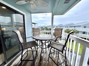 Une terrasse extérieure couverte, avec une table ronde et quatre chaises, un ventilateur de plafond et une vue sur d’autres bâtiments en arrière-plan.