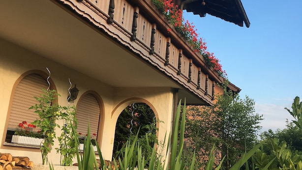 Ein Gebäude mit einem Balkon, der mit roten Blumen geschmückt ist, einer Veranda und einem Garten mit verschiedenen Pflanzen.