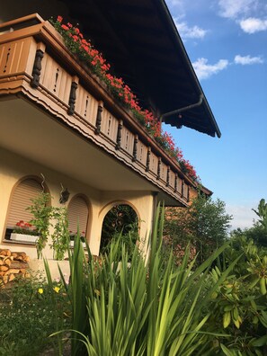 A building with a balcony adorned with red flowers, a covered porch, and a garden with various plants.