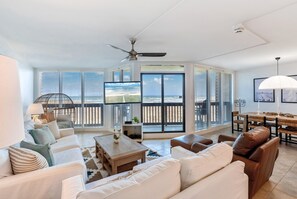 A spacious living room with a large TV, comfortable seating, a balcony with a view, and a ceiling fan.
