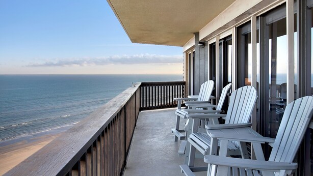 Een balkon met witte Adirondack-stoelen, een houten leuning en uitzicht op de oceaan.