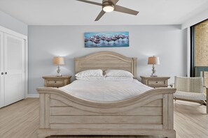 A bedroom with a wooden bed, two bedside tables with lamps, a ceiling fan, a painting, and a wicker chair.