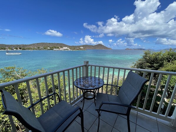 A balcony with a table and two chairs, overlooking a turquoise sea with boats and distant hills.