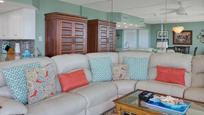 A living room with a sectional sofa, a glass coffee table, and a kitchen in the background. - Sandpiper Condominiums 107L, Panoramic Gulf Views w/ direct access to Mustang Island/Port A beaches. (Port Aransas)