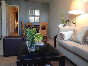 Een woonkamer met een glazen salontafel, een bank, een lamp en ingelijste foto’s aan de muur.